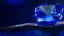Blue Sapphire gemstone, cut in a cushion or emerald shape, resting on a dark textured surface under dramatic blue lighting.